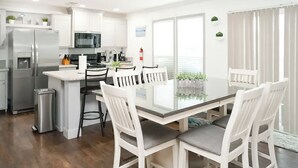 Una cocina moderna con armarios blancos, electrodomésticos de acero inoxidable y un comedor con mesa de vidrio y sillas blancas.