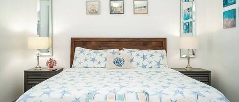 A bedroom with a bed featuring a 'Beach House' pillow, star-patterned bedding, and framed pictures on the wall.