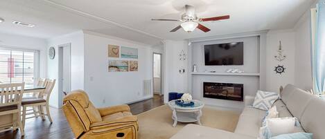 A modern living room with a beige sofa, a tan recliner, and a white couch. A flat-screen TV is mounted above a fireplace.
