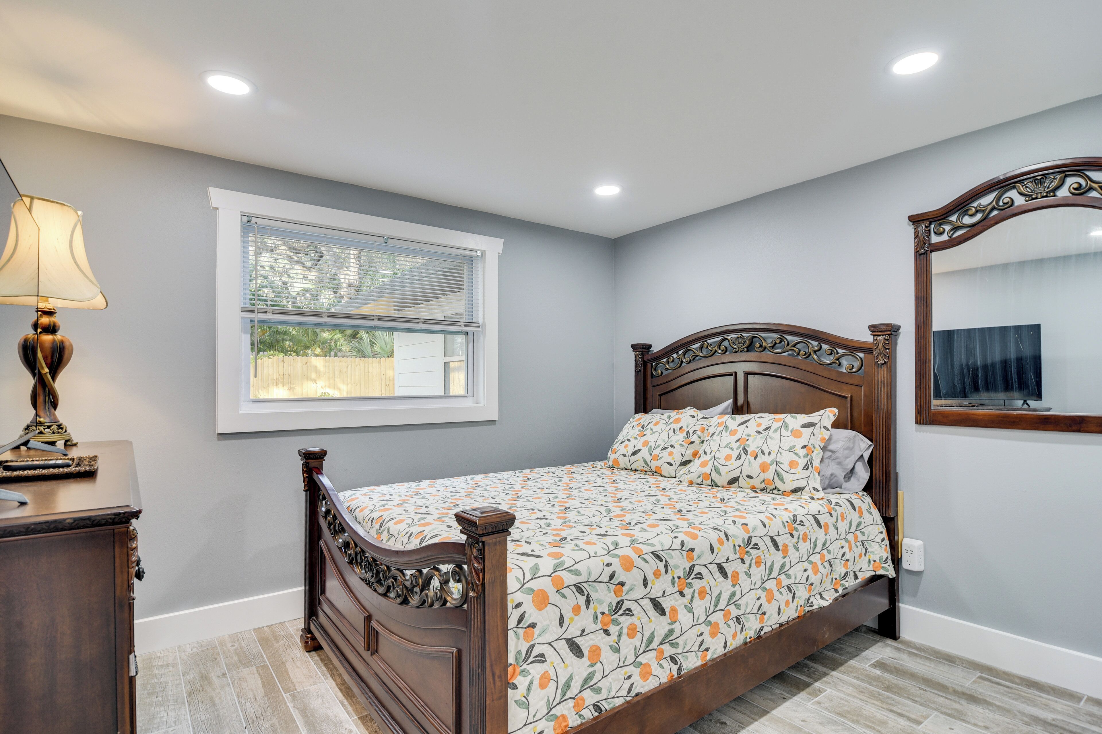 Une chambre à coucher avec un lit en bois, une table de chevet, une lampe, une fenêtre avec des stores et un miroir sur le mur.