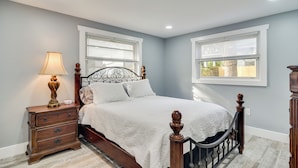 Un dormitorio con cama, mesita de noche, lámpara y dos ventanas.