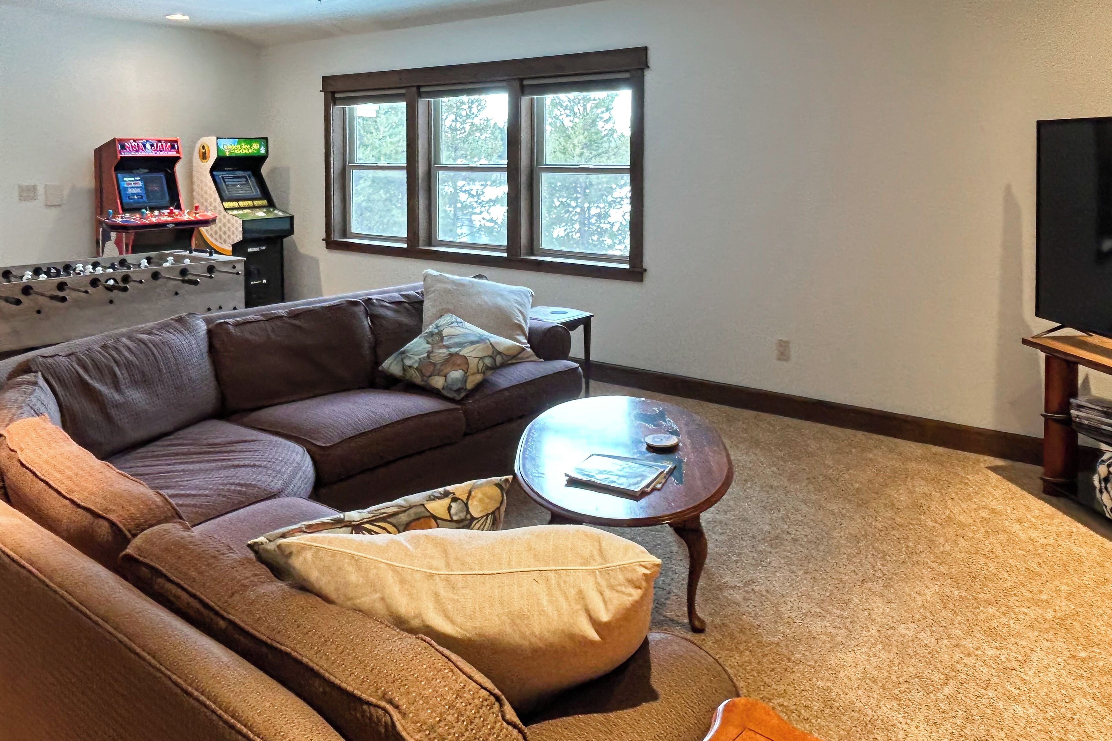 A living room with a brown sofa, a coffee table, a foosball table, and two arcade machines.