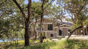 A two-story house with a large deck, surrounded by trees and a body of water. - Lakefront, Private 3br, Dog-friendly Rental (Canyon Lake)