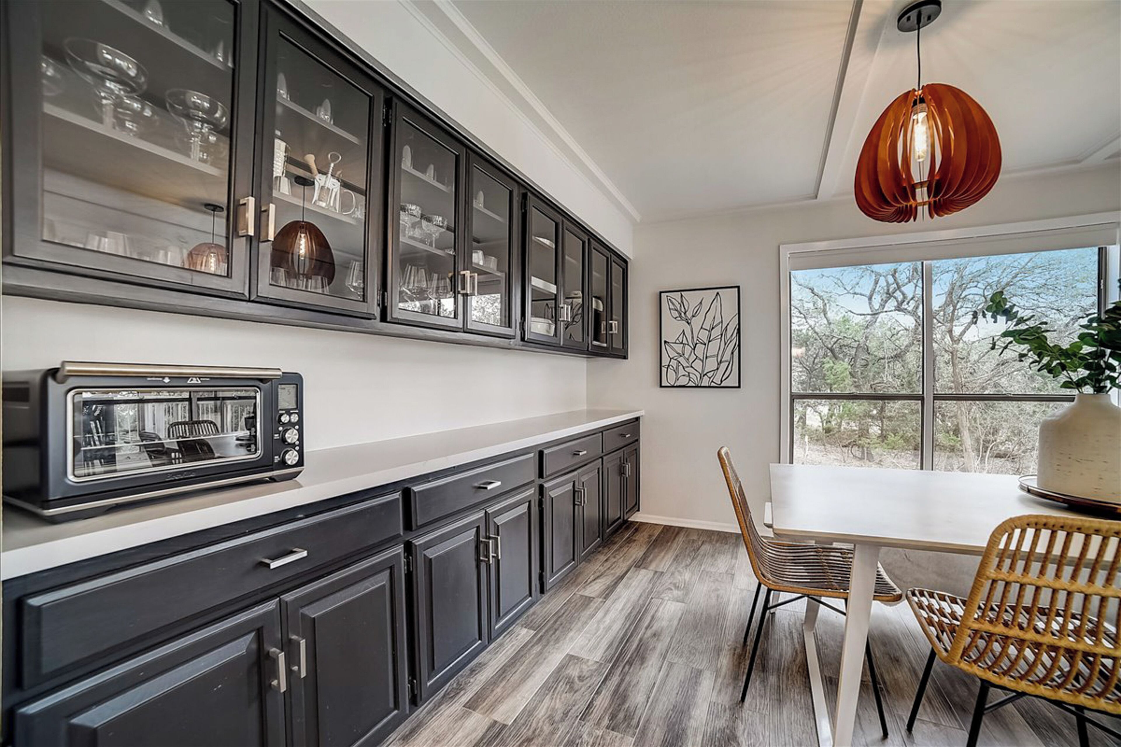 A modern kitchen with dark cabinets, a built-in microwave, and a dining area with wicker chairs and a large window.
