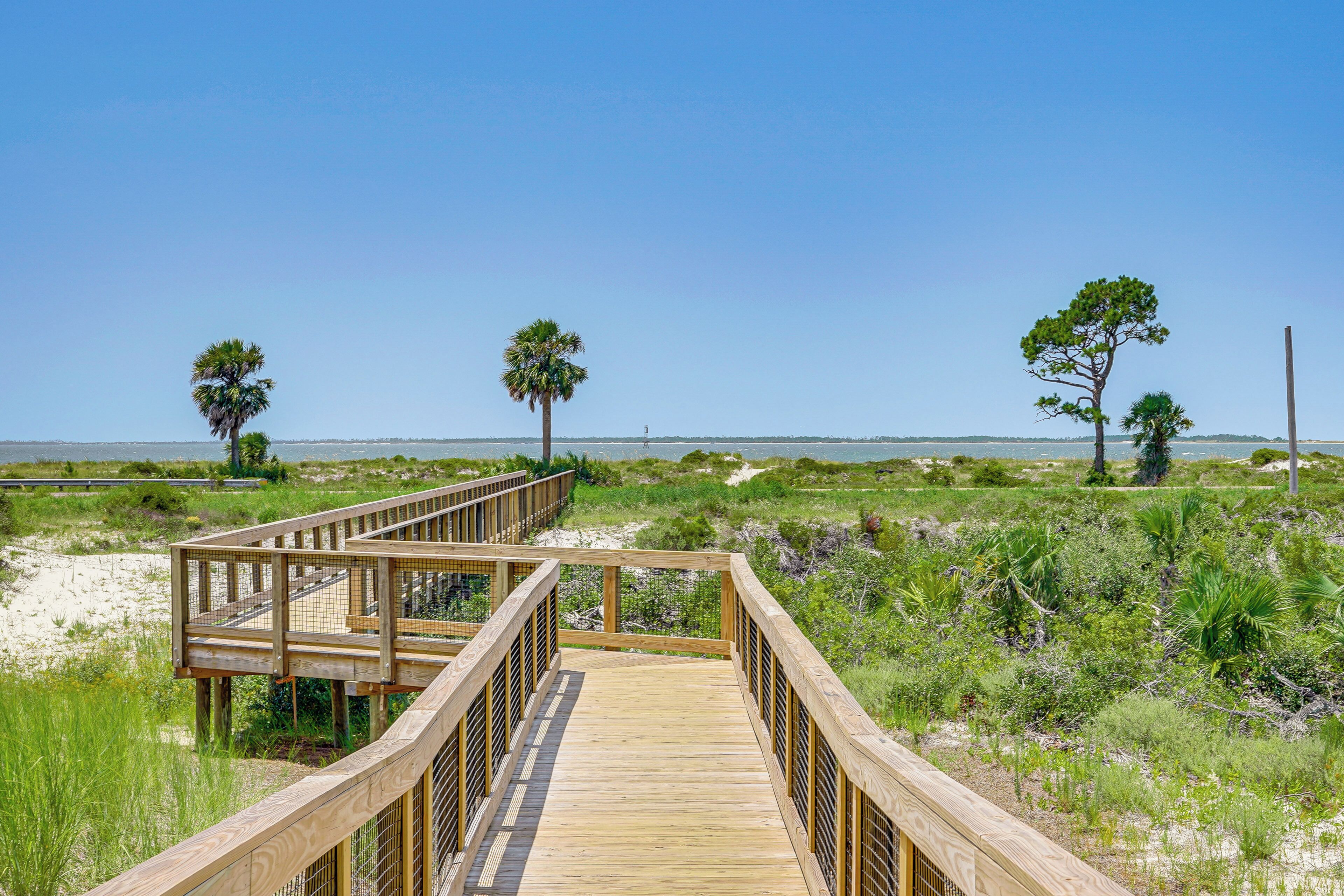 A wooden boardwalk leading to a beach with sparse vegetation and a clear sky.