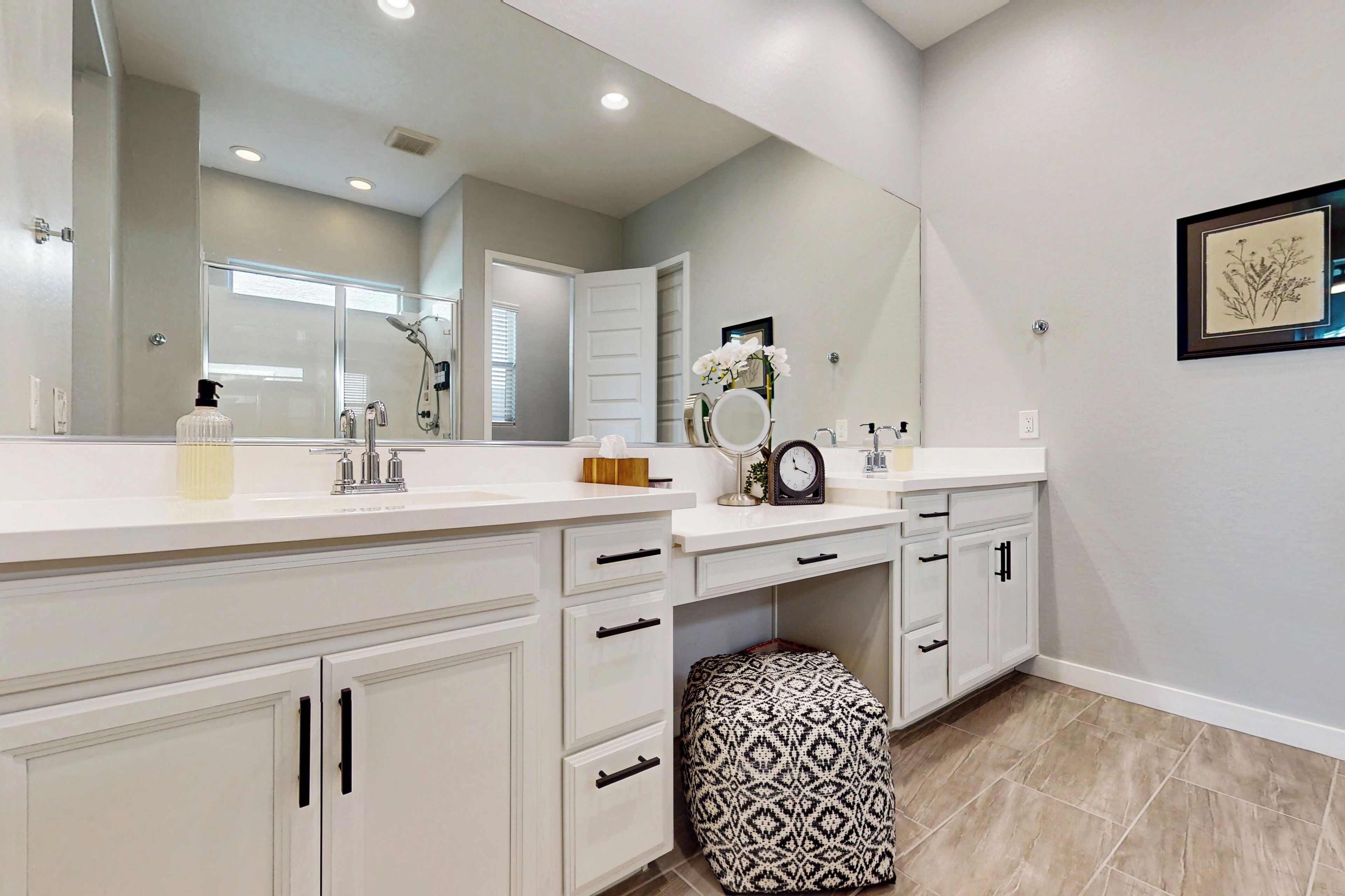 Un baño moderno con un espejo grande, muebles blancos, un otomano estampado y una obra de arte enmarcada en la pared.
