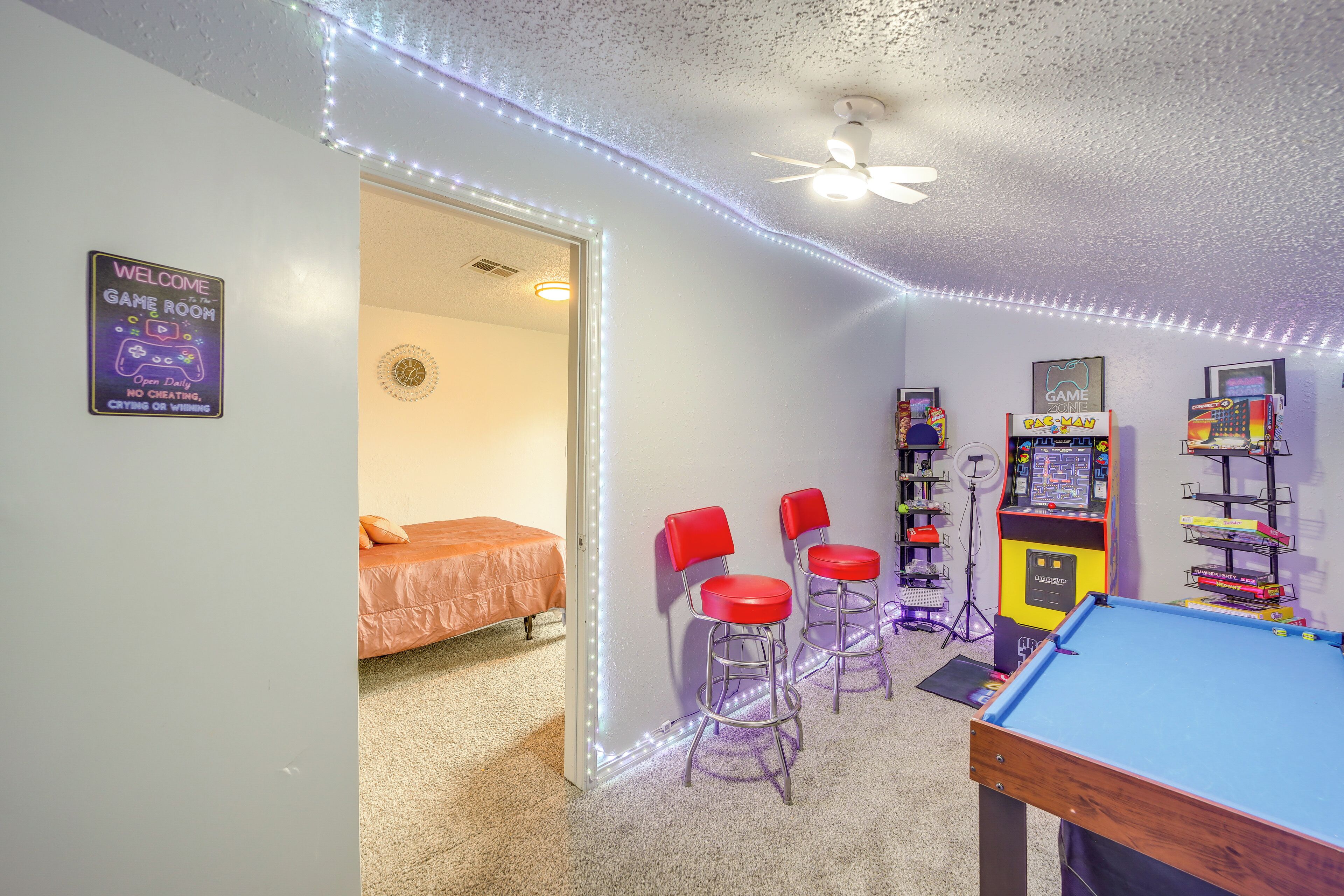 Ein Raum mit Billardtisch, zwei roten Barhocker, Deckenventilator und Spielautomaten.