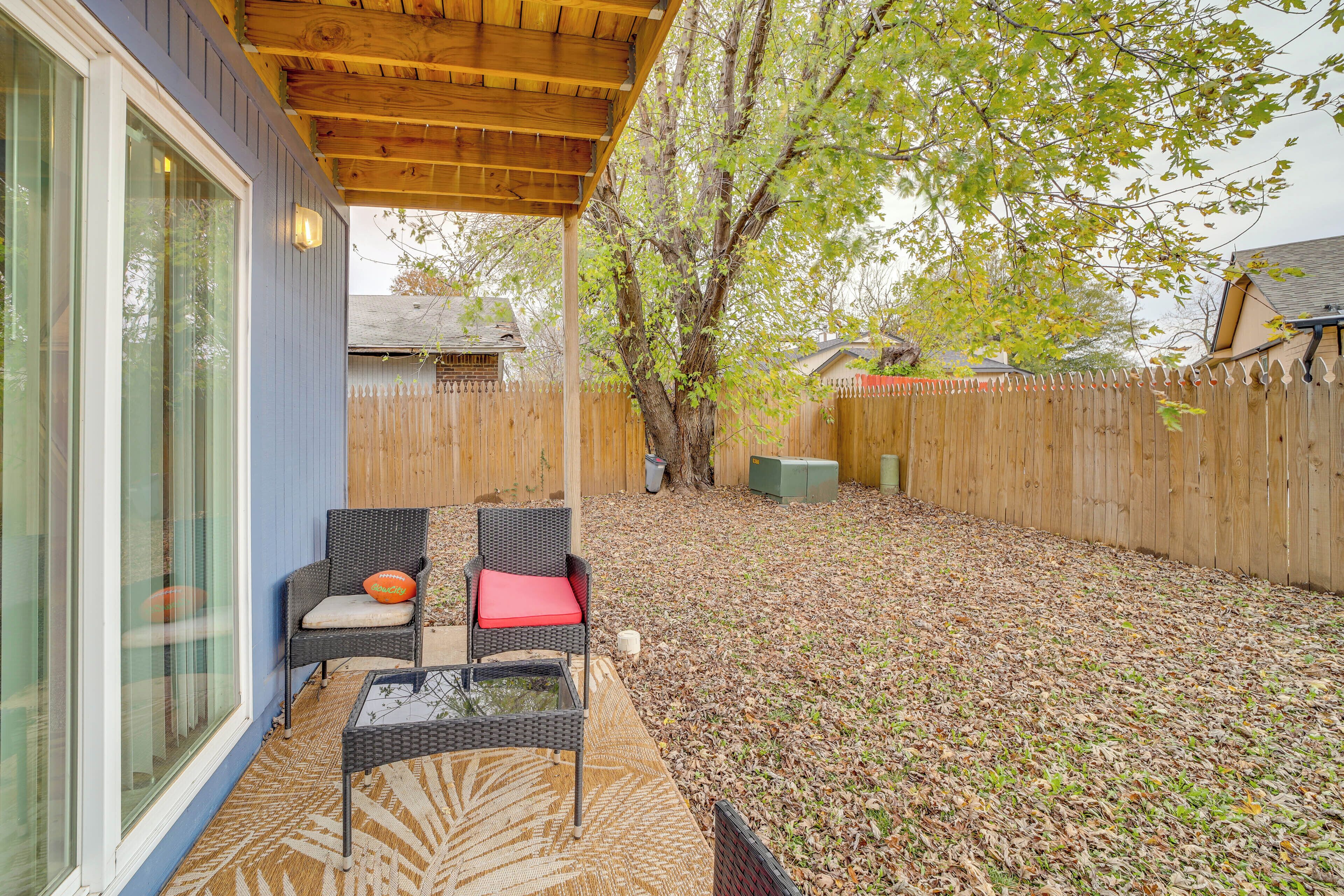 A covered patio area with two wicker chairs and a glass table, a wooden fence, and a tree in the background.