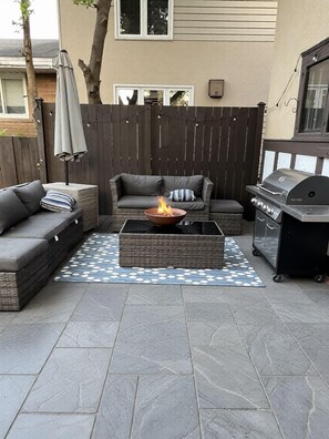 An outdoor patio with a gray sofa, a fire pit, and a grill.
