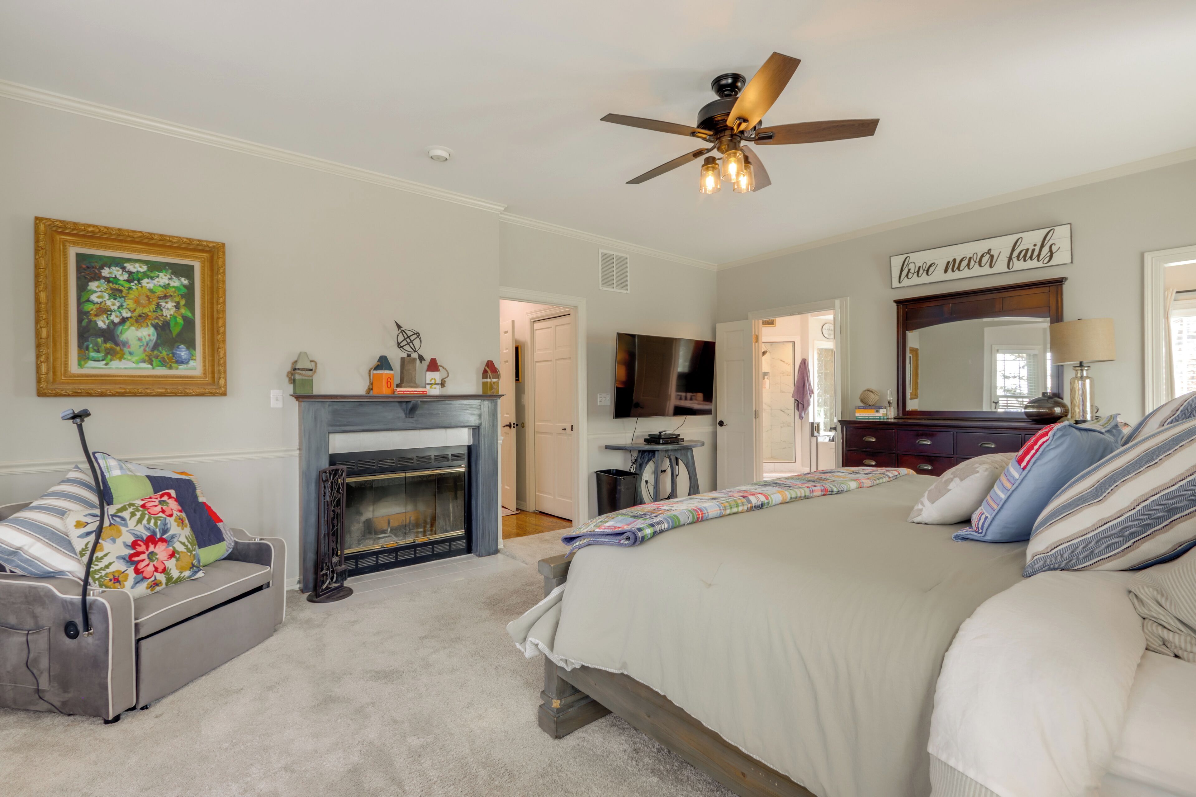 A bedroom with a fireplace, a ceiling fan, a bed with a quilt, a sofa, and a painting on the wall.