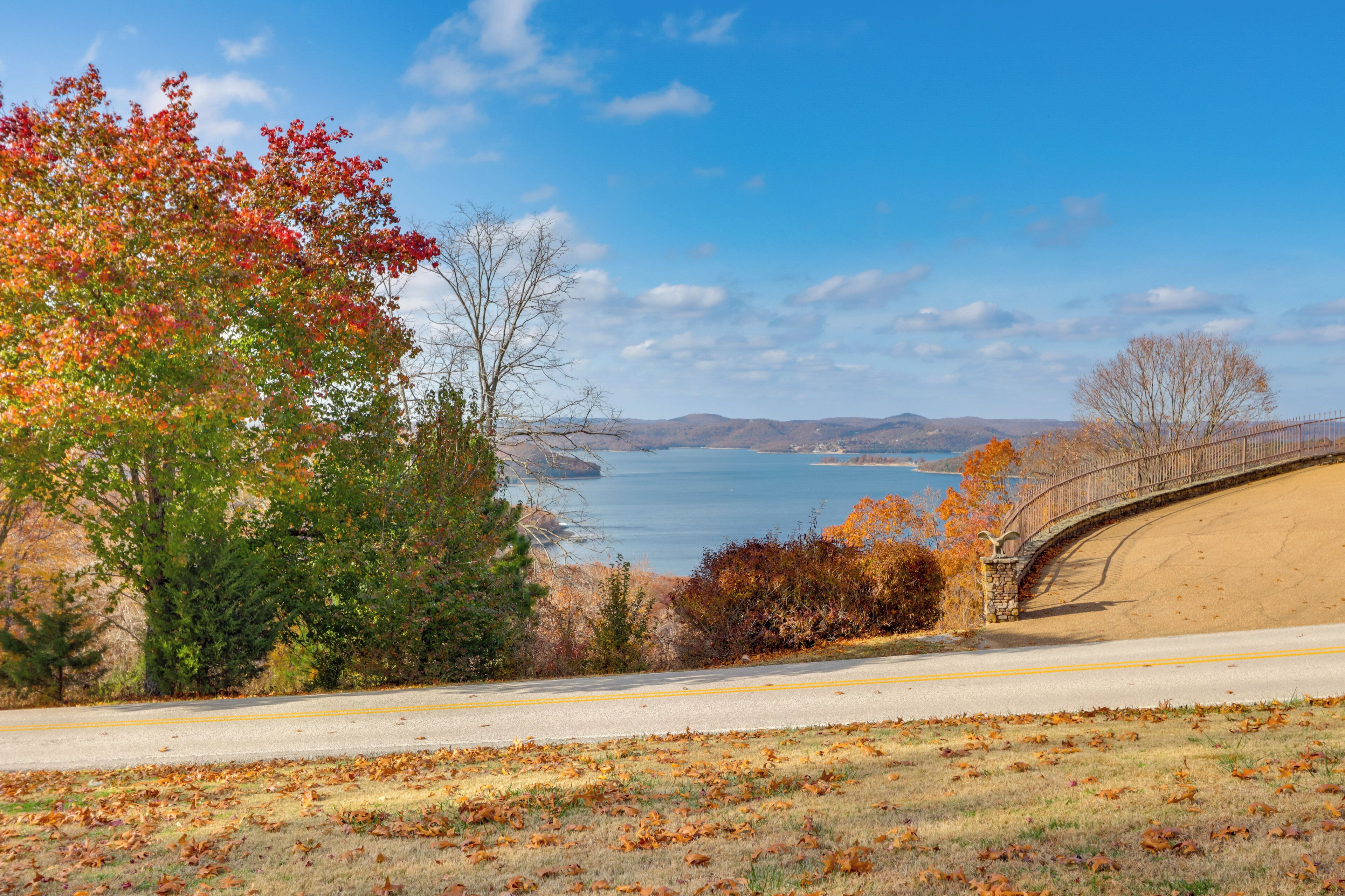 A scenic view of a coastal road with a stone bridge, autumn foliage, and a clear blue sky.