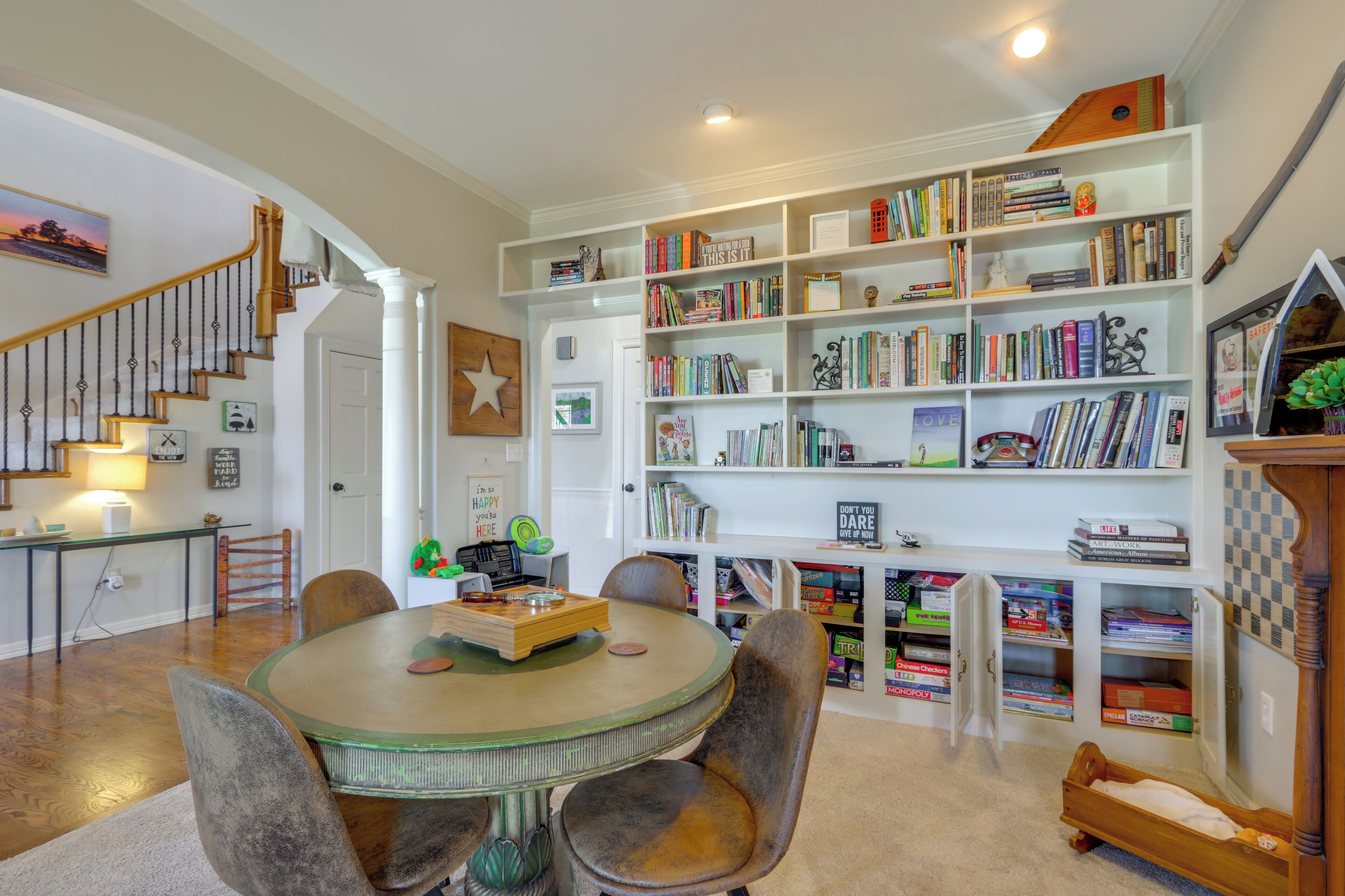 A spacious living room with a wooden staircase, a round dining table, and a large bookshelf filled with books.
