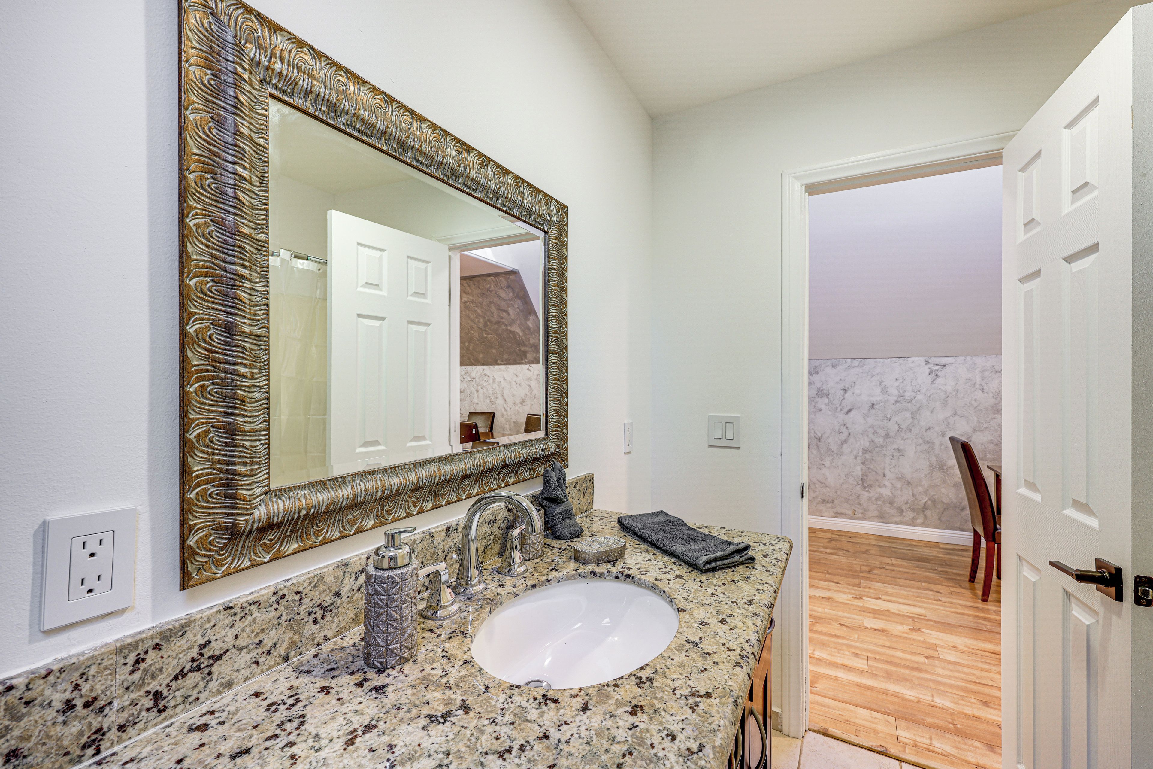 A bathroom with a granite countertop, a white sink, a large mirror with an ornate frame, and a door leading to another room.