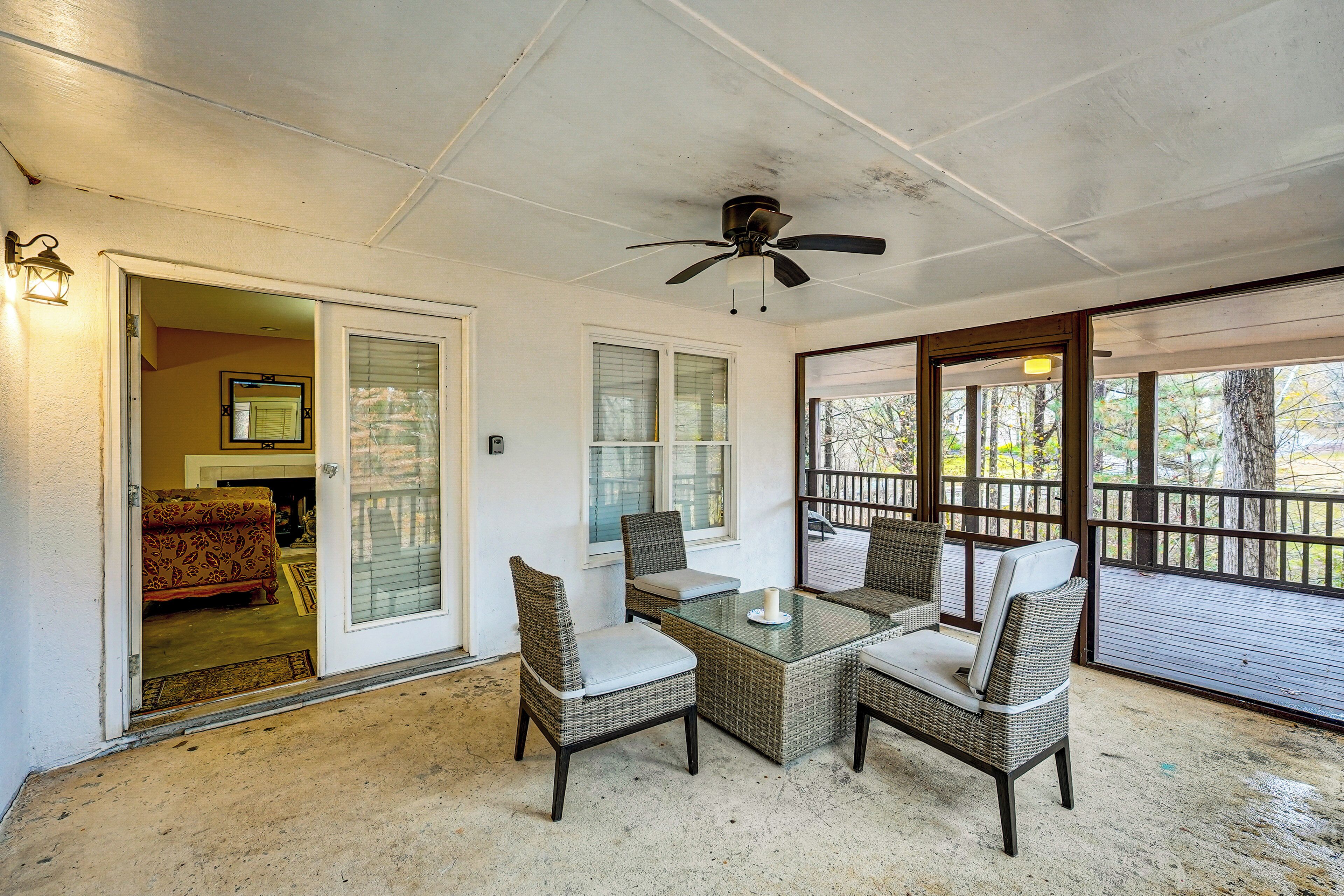 A covered patio area with a glass table and wicker chairs, a ceiling fan, and a view of the outdoors through large windows.
