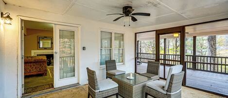 A covered patio area with a glass table and wicker chairs, a ceiling fan, and a view of the outdoors through large windows.