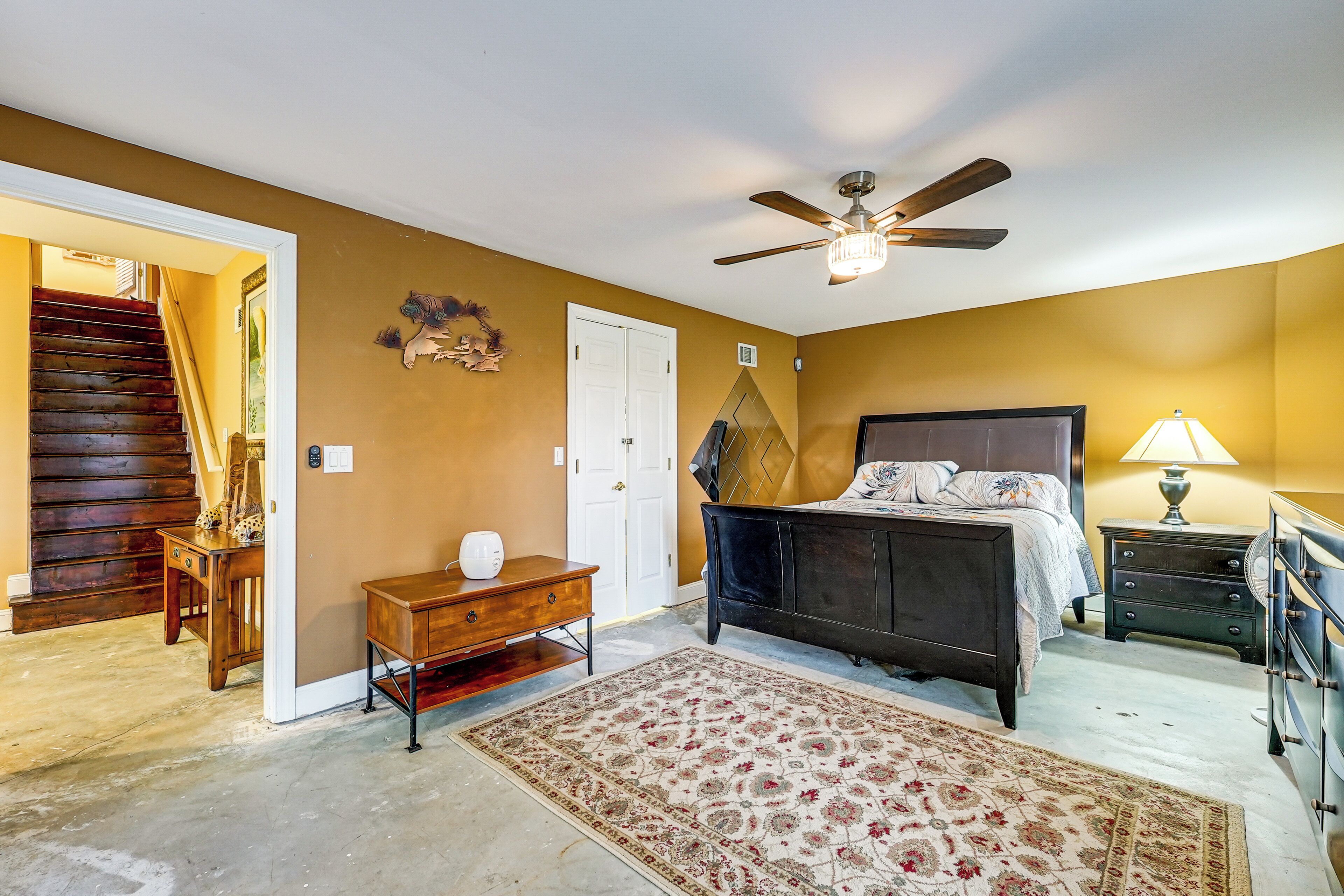 Une chambre à coucher comprenant un lit, un ventilateur de plafond, une table de chevet, une commode, un tapis et un escalier menant à une autre pièce.