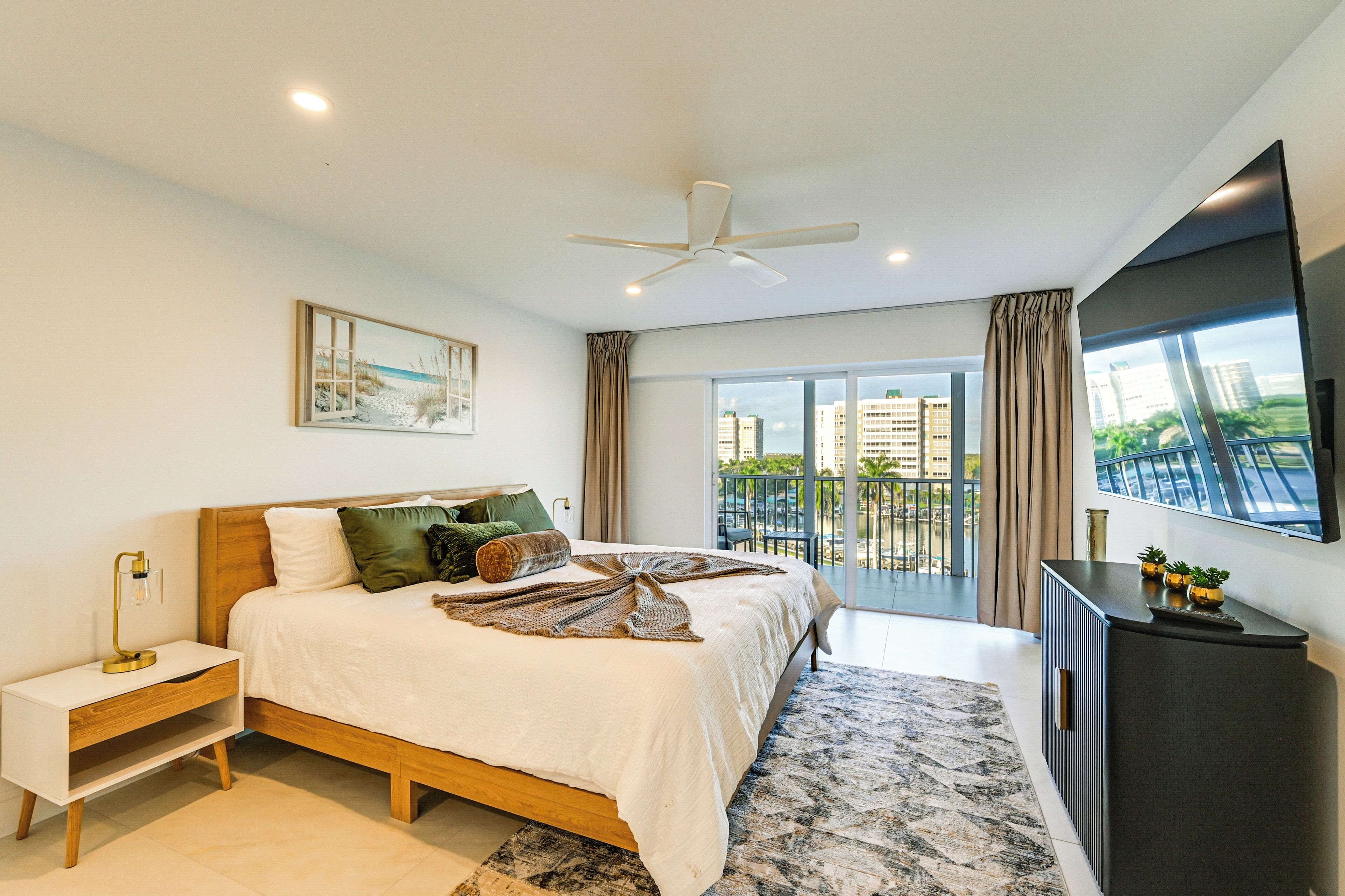 A modern bedroom with a bed, bedside table, and a large window with a view of buildings.