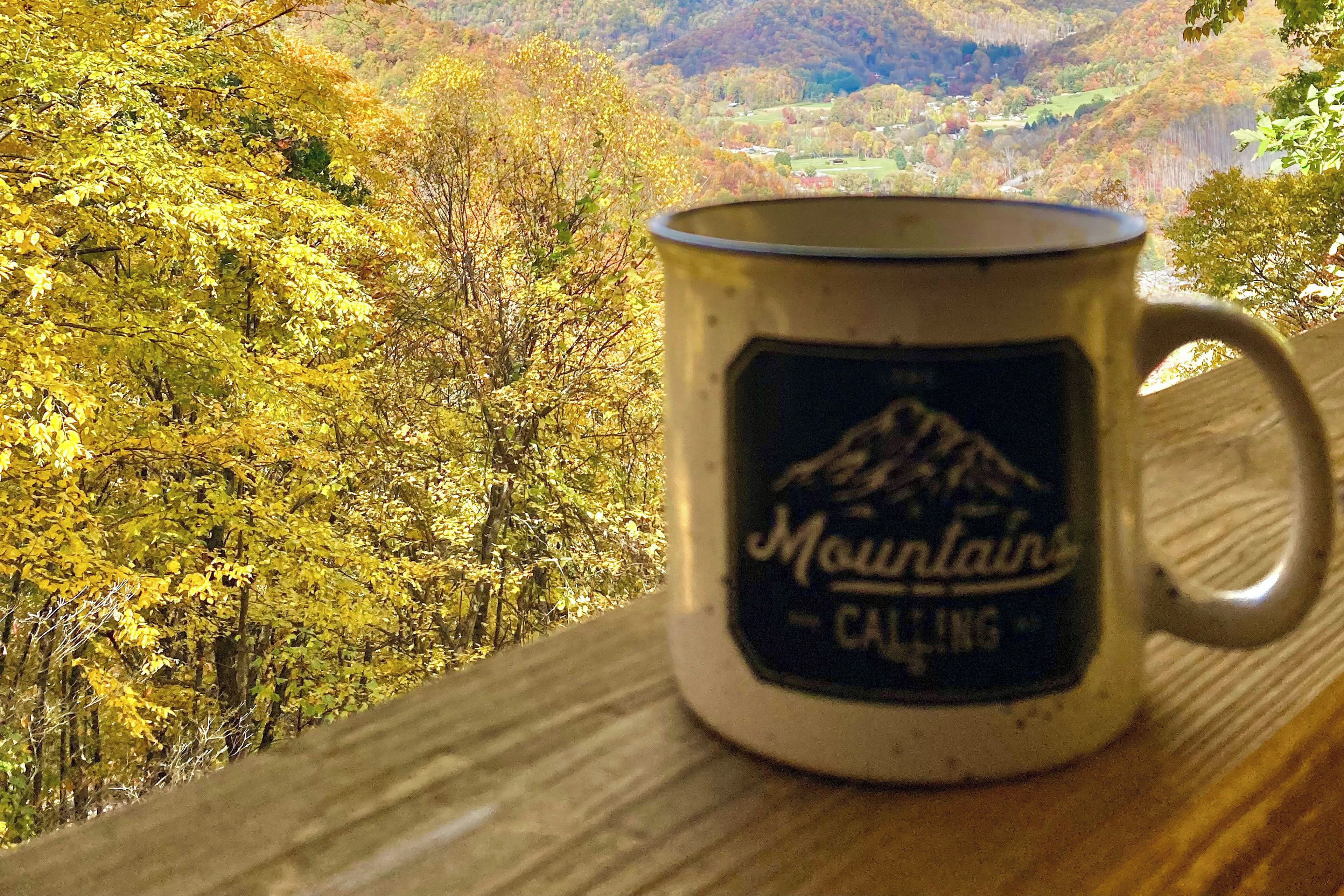 A mug with a mountain design on a wooden surface with a scenic view of autumn trees in the background.