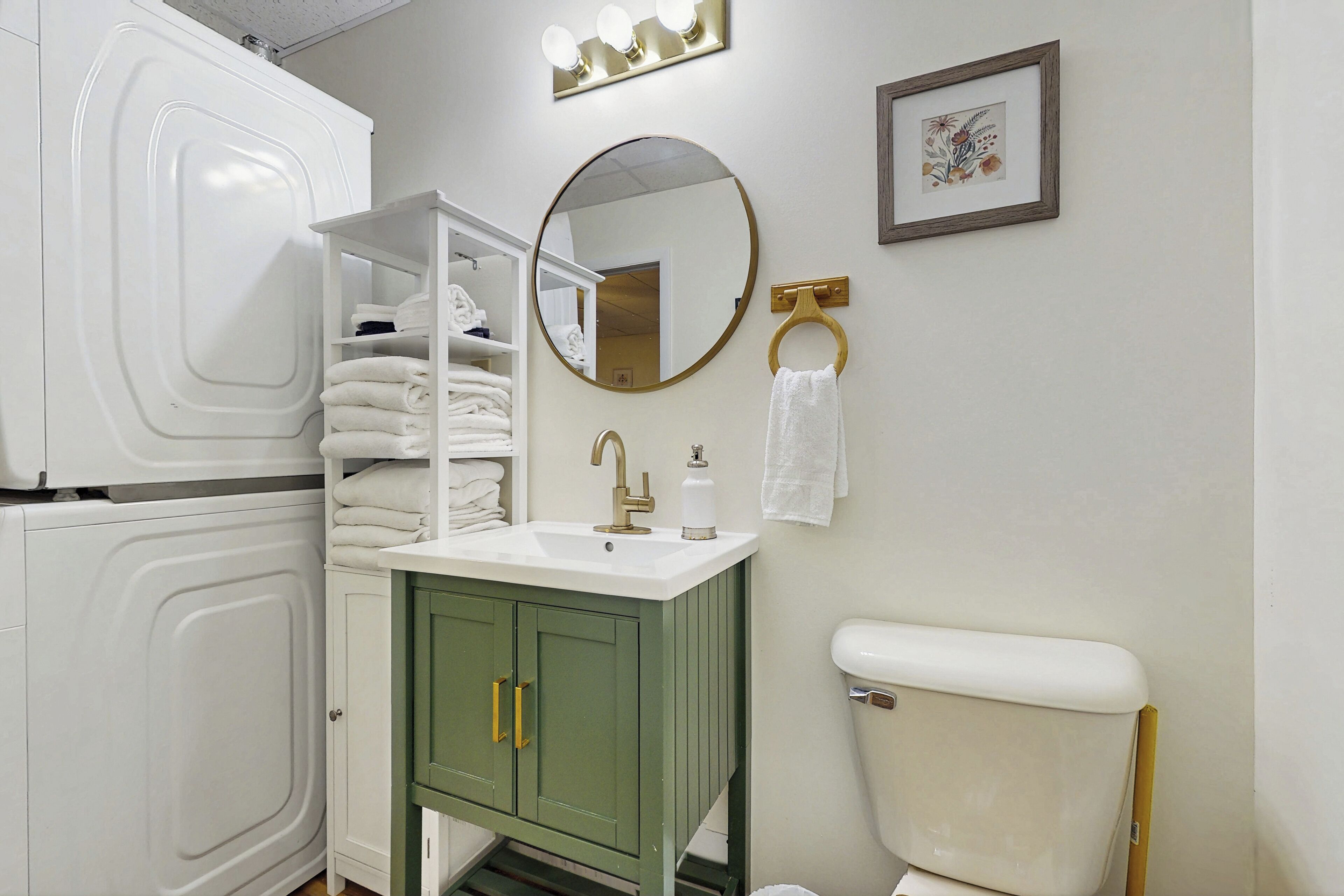 Une salle de bain avec des toilettes blanches, un meuble-lavabo vert avec des poignées dorées, un lavabo blanc et un miroir avec des lumières au-dessus.