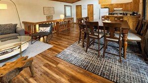 Amplia sala de estar con piso de madera, una mesa de comedor con sillas, un sofá y una cocina con gabinetes de madera y una lámpara colgante.