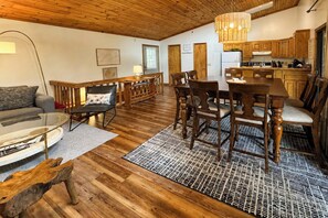 Amplia sala de estar con piso de madera, una mesa de comedor con sillas, un sofá y una cocina con gabinetes de madera y una lámpara colgante.