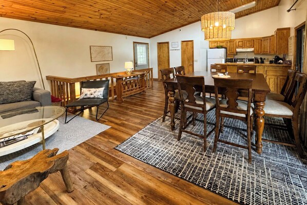 Uma sala ampla com piso de madeira, uma mesa de jantar com cadeiras, um sofá e uma área de cozinha com armários de madeira e uma luminária pendente.