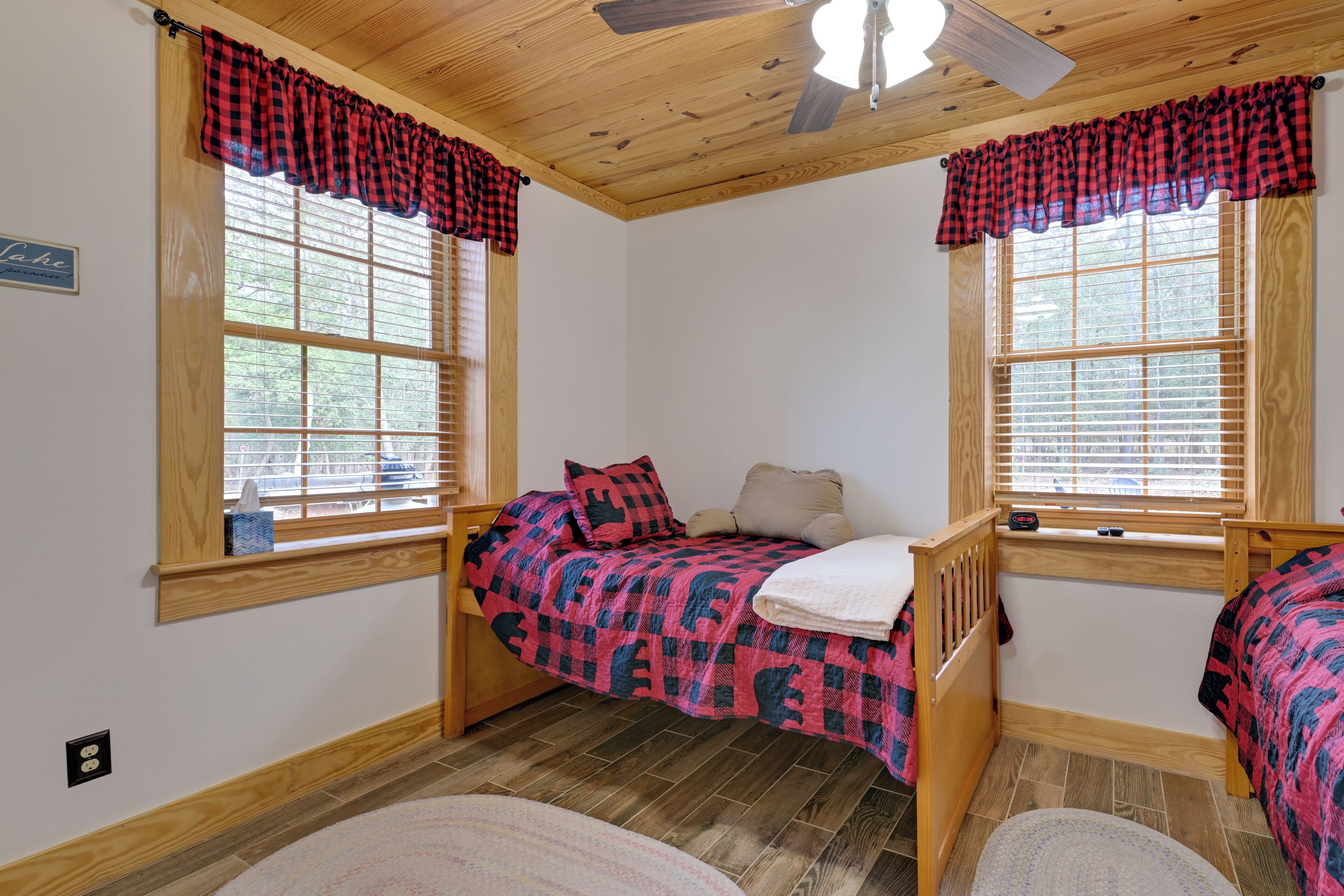 A bedroom with two beds, wooden flooring, a ceiling fan, and large windows with blinds.