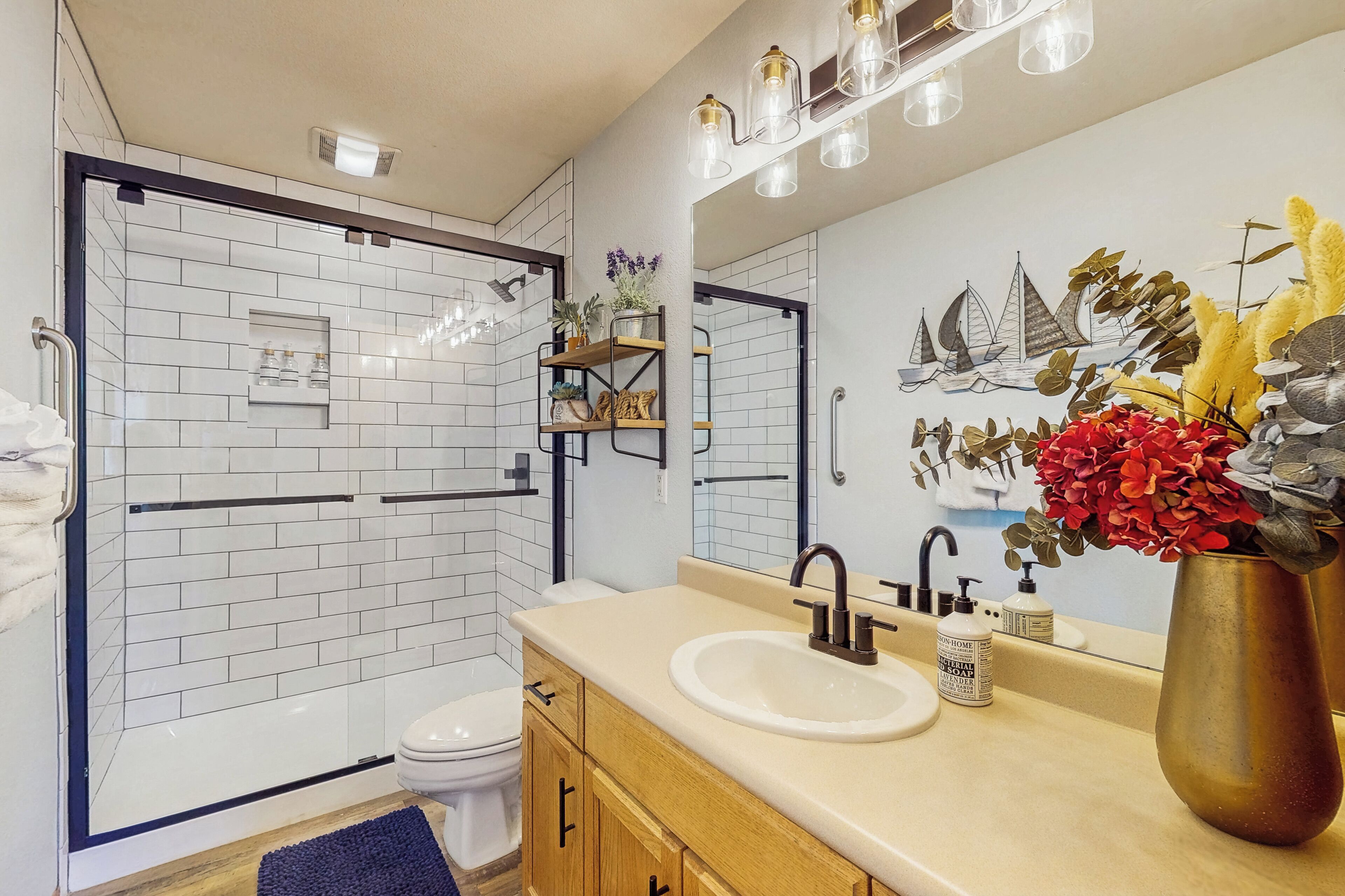 Een moderne badkamer met een glazen douchecabine, een wit toilet, een beige wastafel met een gouden vaas met bloemen en een wandspiegel met een nautisch muurschildering.