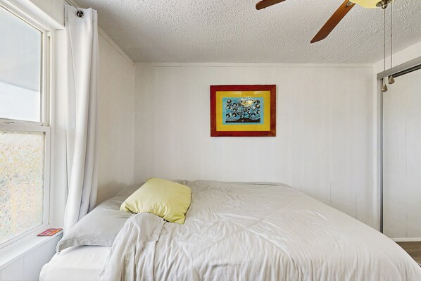 Une chambre à coucher avec un lit, une fenêtre avec des rideaux, un ventilateur de plafond et un tableau encadré au mur.