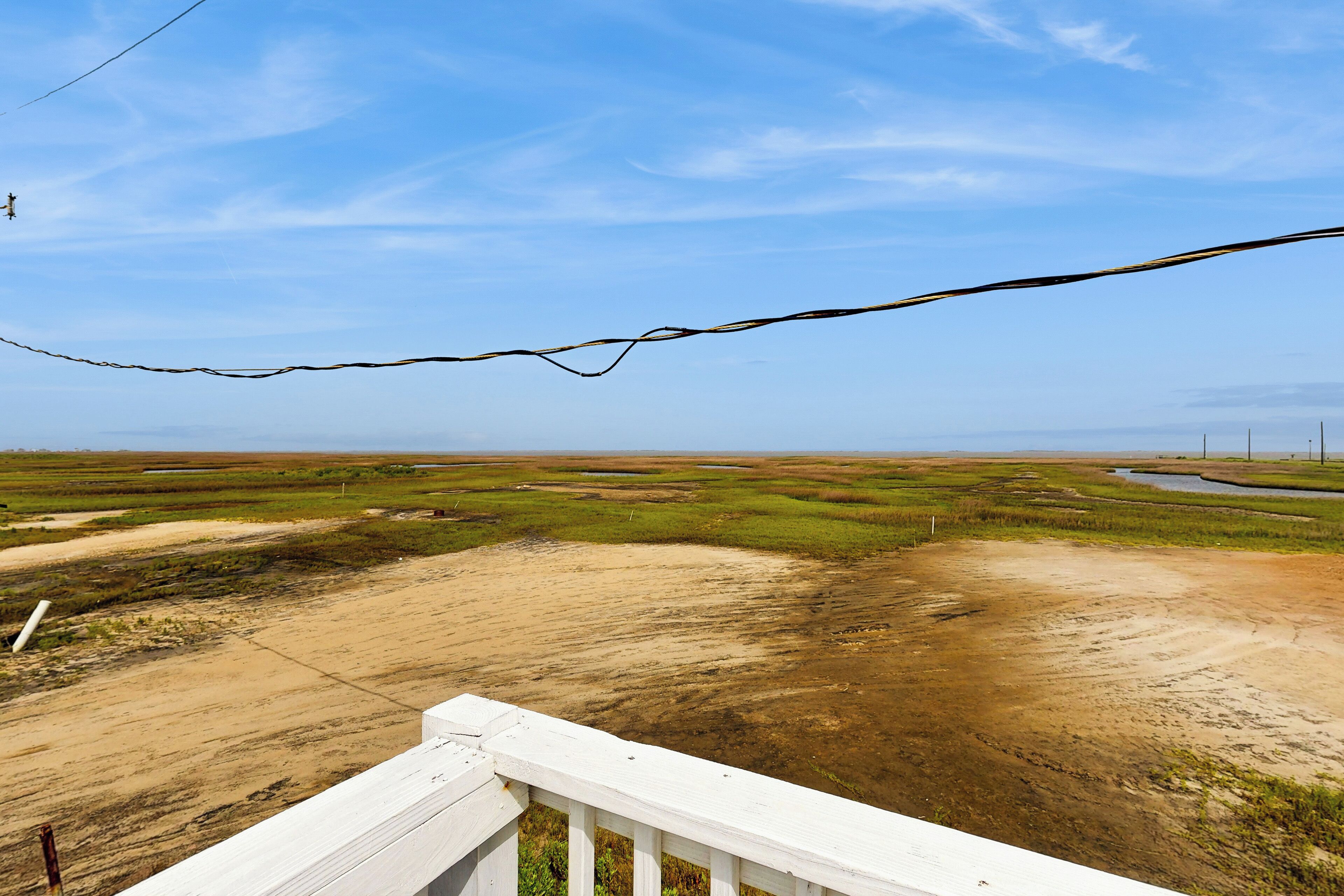 A view from a balcony with a railing, overlooking a landscape with patches of grass and dirt, and a clear blue sky.