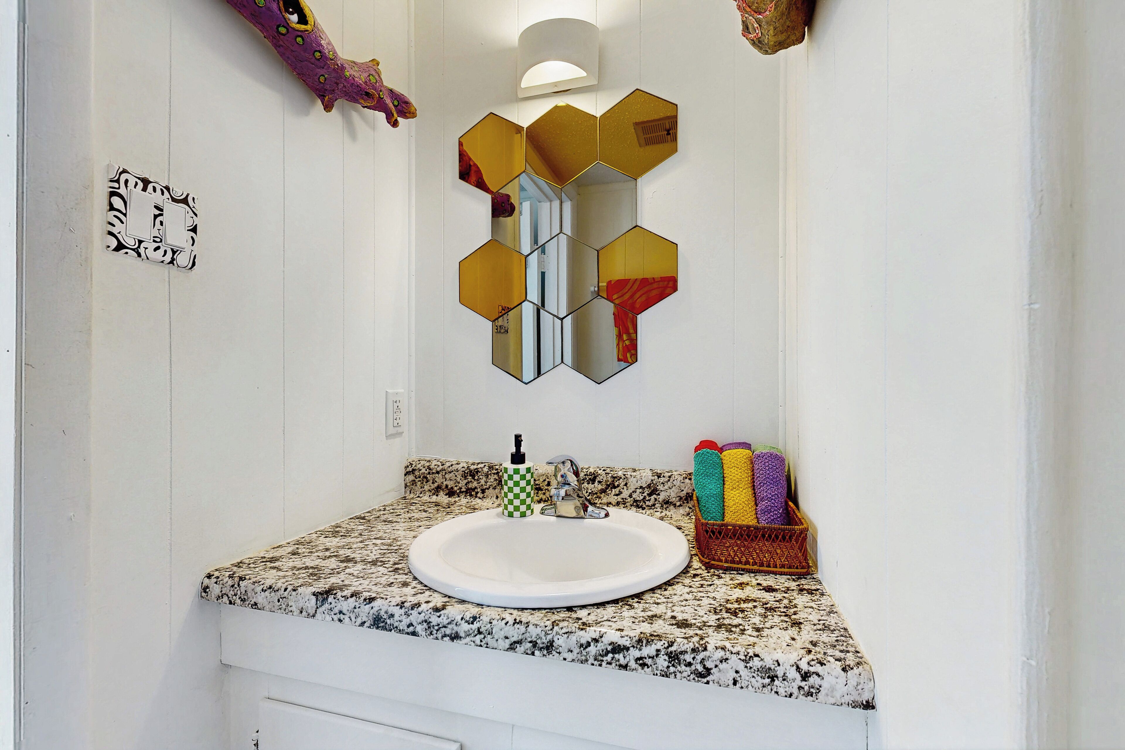 A bathroom with a granite countertop, a white sink, a mirror with hexagonal frames, and a towel rack with colorful towels.