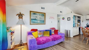 Un salón vibrante con un sofá morado, cojines de colores, un cuadro enmarcado de un paisaje y una cortina color turquesa.