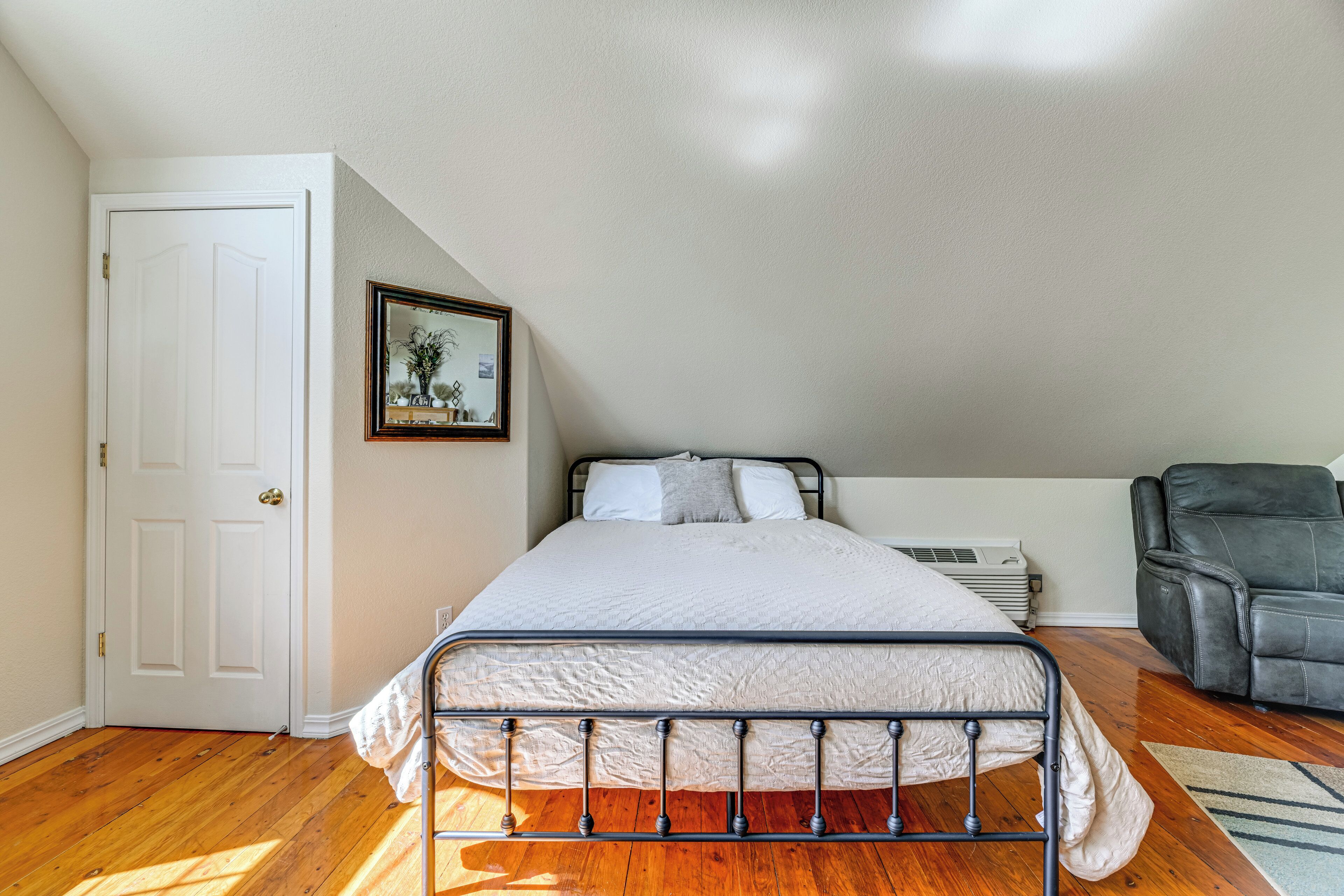 Un dormitorio con cama, puerta, cuadro enmarco, silla y calefactor.