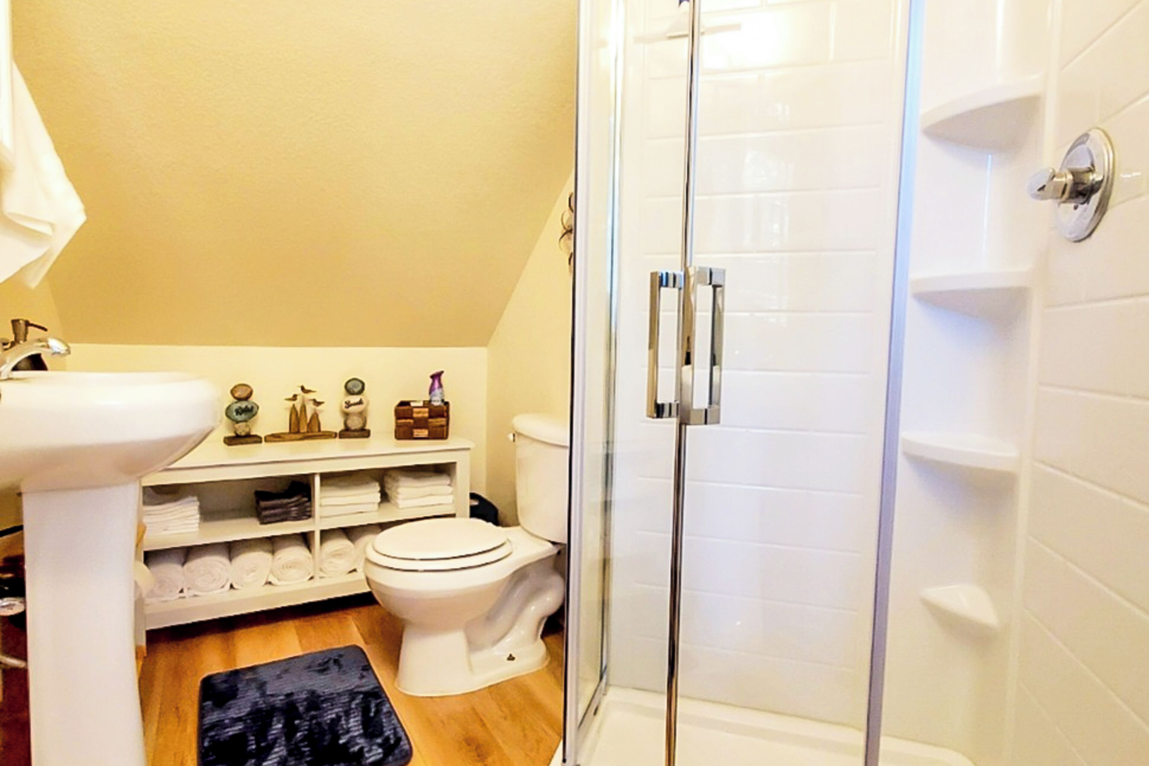 A bathroom with a glass-enclosed shower, a white sink, a toilet, and a shelf with towels and toiletries.