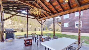 Un porche techado al aire libre con una mesa y sillas, una parrilla de barbacoa y vistas a una casa de madera.
