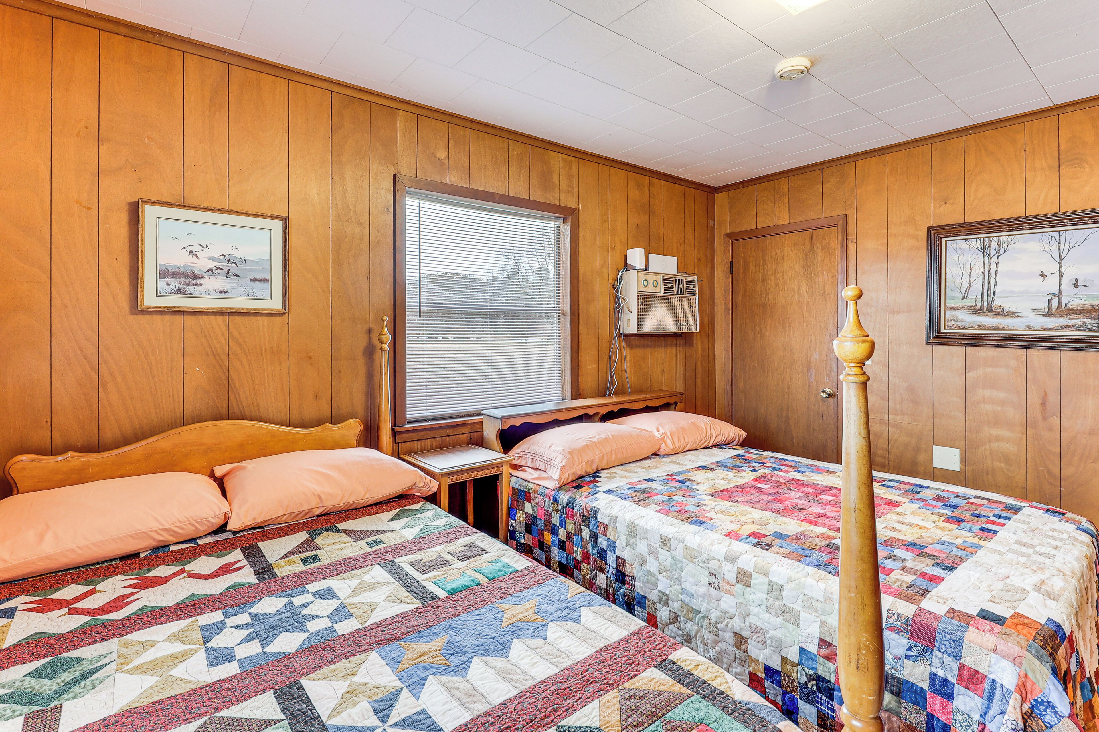 A room with two beds, wooden walls, a window with blinds, a heater, and framed pictures on the walls.