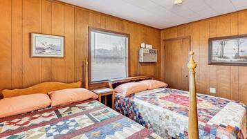 A room with two beds, wooden walls, a window with blinds, a heater, and framed pictures on the walls.