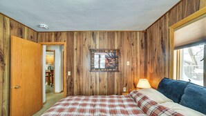 A bedroom with wooden paneling, a plaid bedspread, a nightstand with a lamp, and a window with blinds. - Spacious Retreat w/ Private Yard & Deck in Roscoe! (Roscoe)