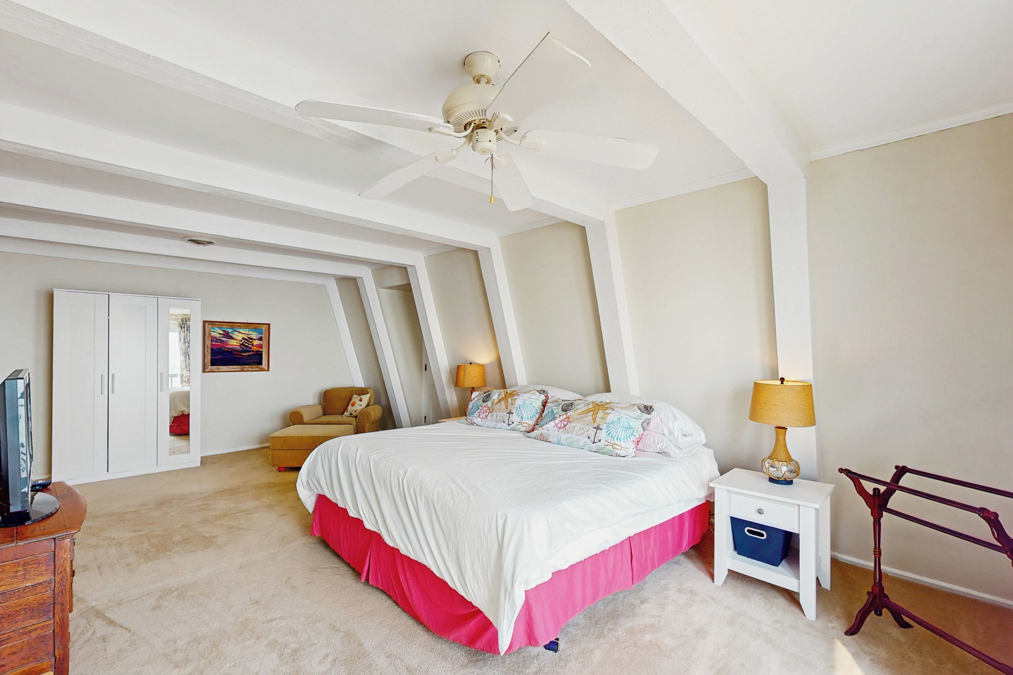 A bedroom with a large bed, a ceiling fan, a nightstand with a lamp, a wardrobe, and a painting on the wall.