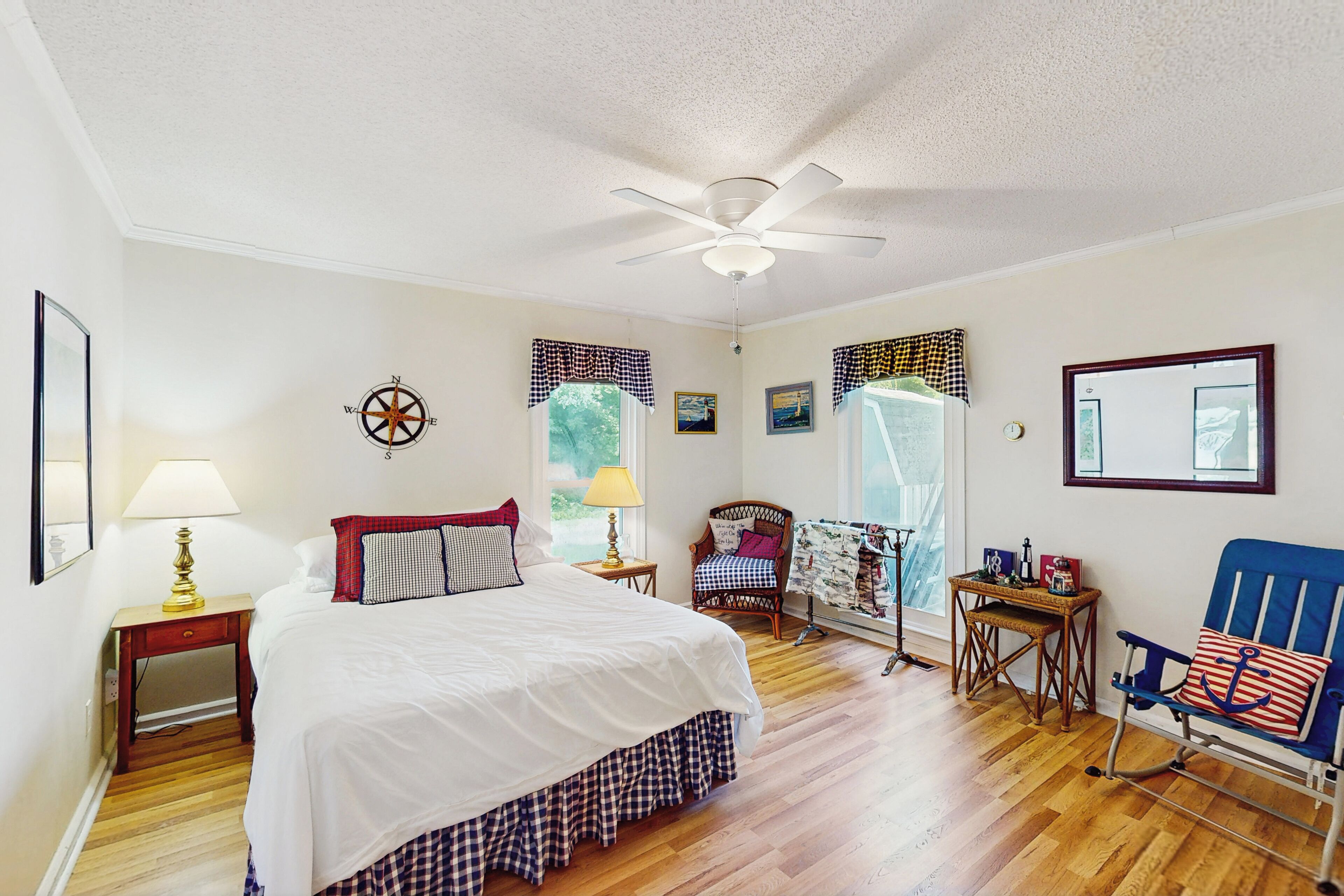 Une chambre avec un grand lit, un ventilateur de plafond, une fenêtre avec des rideaux, un fauteuil à bascule et une petite table avec une lampe.