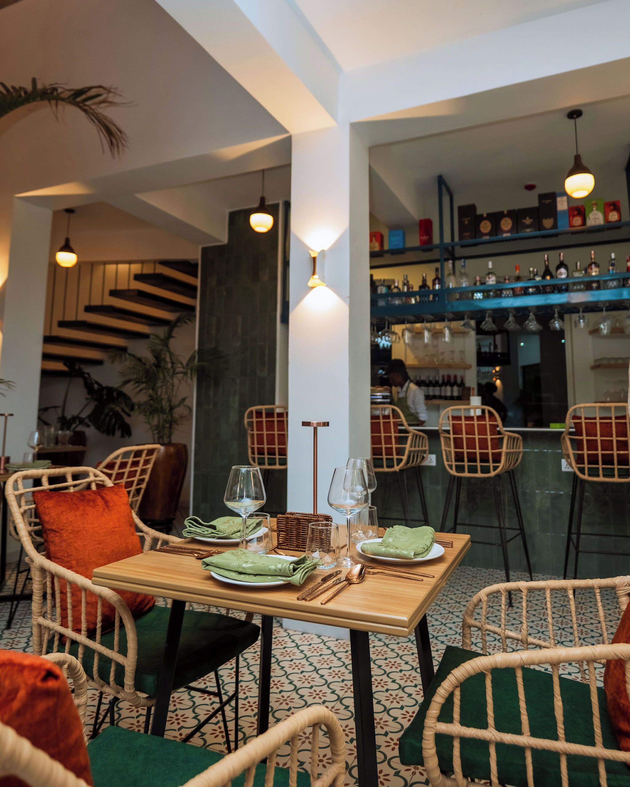 Un coin repas avec des chaises en osier, une table en bois dressée avec des assiettes, des verres et des couverts. On aperçoit des lampes suspendues et un bar au fond.