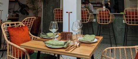 Un comedor con sillas de mimbre, una mesa de madera dispuesta con platos, vasos y cubiertos. Se observan luces colgantes y una zona de bar al fondo.