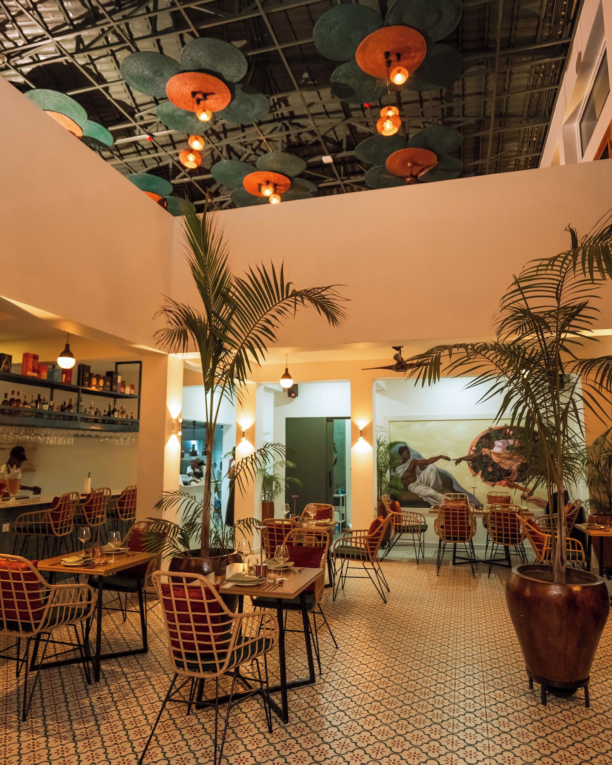 A restaurant with high ceilings, hanging pendant lights, and a patterned floor. Tables are set with chairs, and there are potted plants.