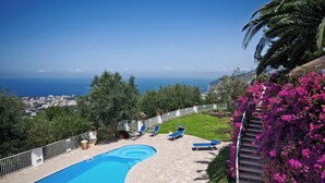 Una piscina con vistas al mar, rodeada de un jardín con flores rosadas y una palmera.
