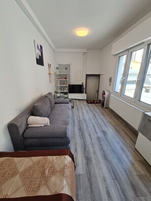 A modern living room with a grey sofa, a flat-screen TV, a wooden floor, and a window with blinds. - Cityapartment +terrace up to 4 persons Neuwied city center Rheinprominade (Neuwied)
