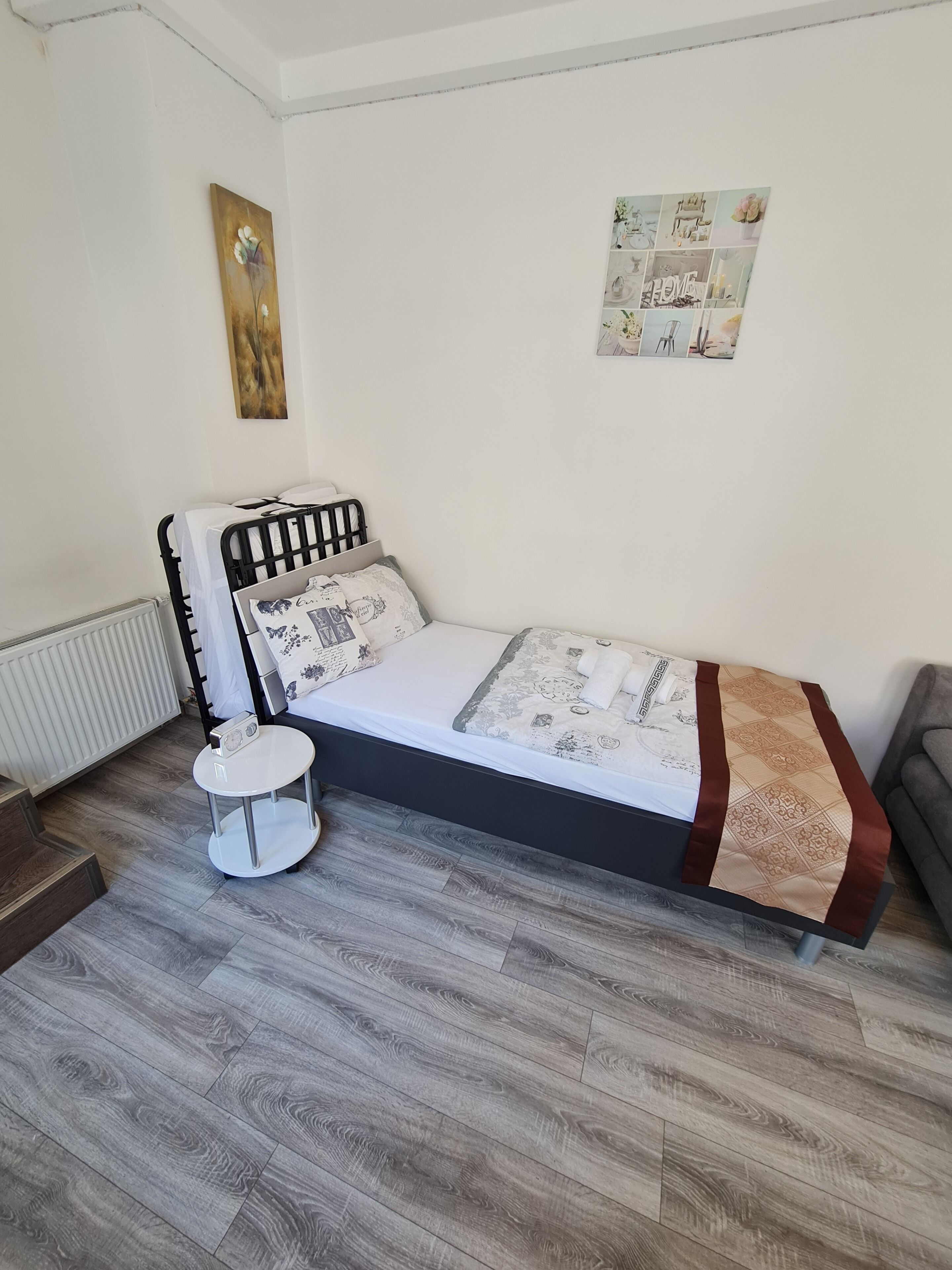 Een slaapkamer met een bed, nachtkastje, radiator en een muur met foto's.