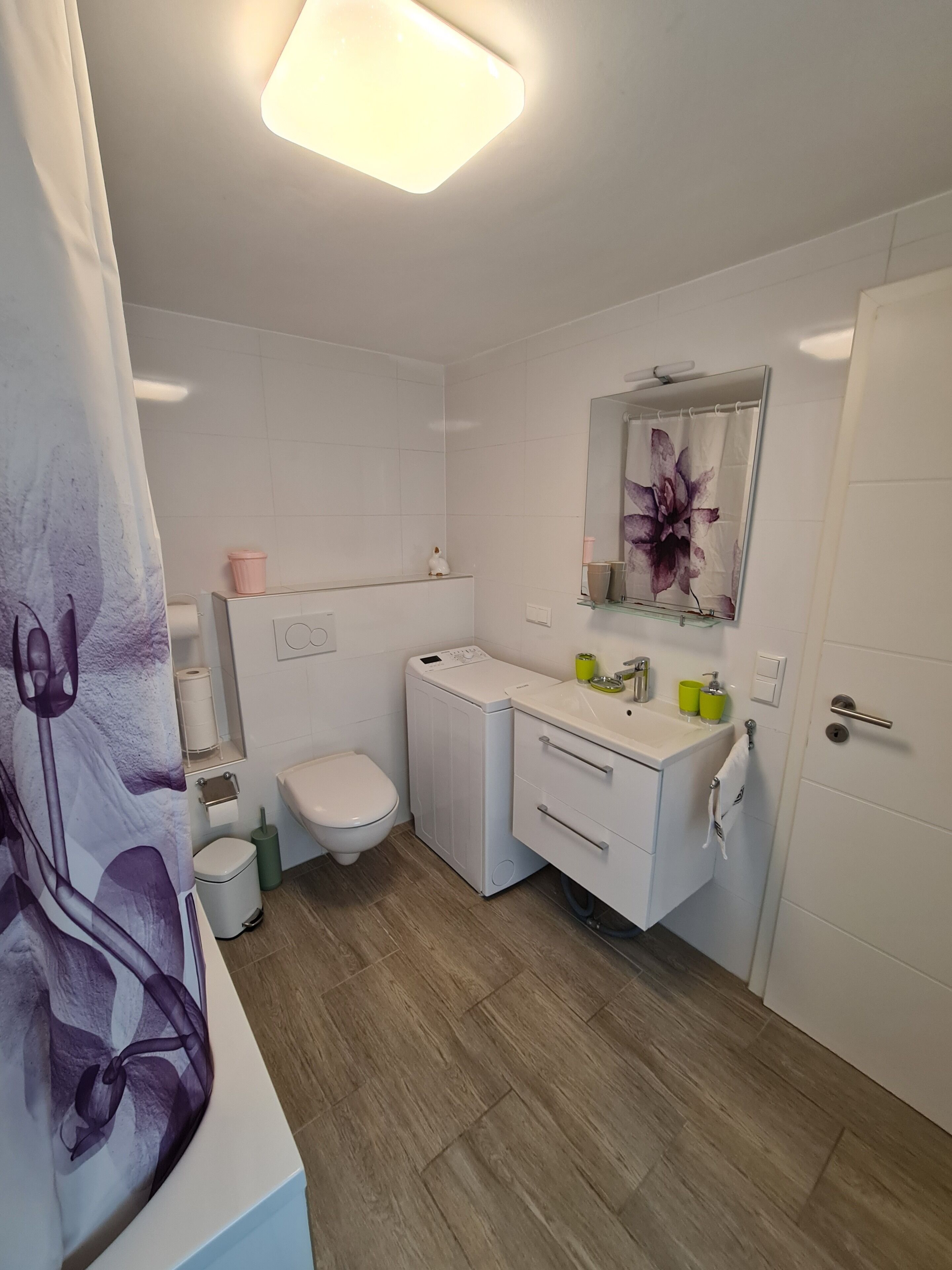 A bathroom with a toilet, washing machine, sink, mirror, and a shower curtain.