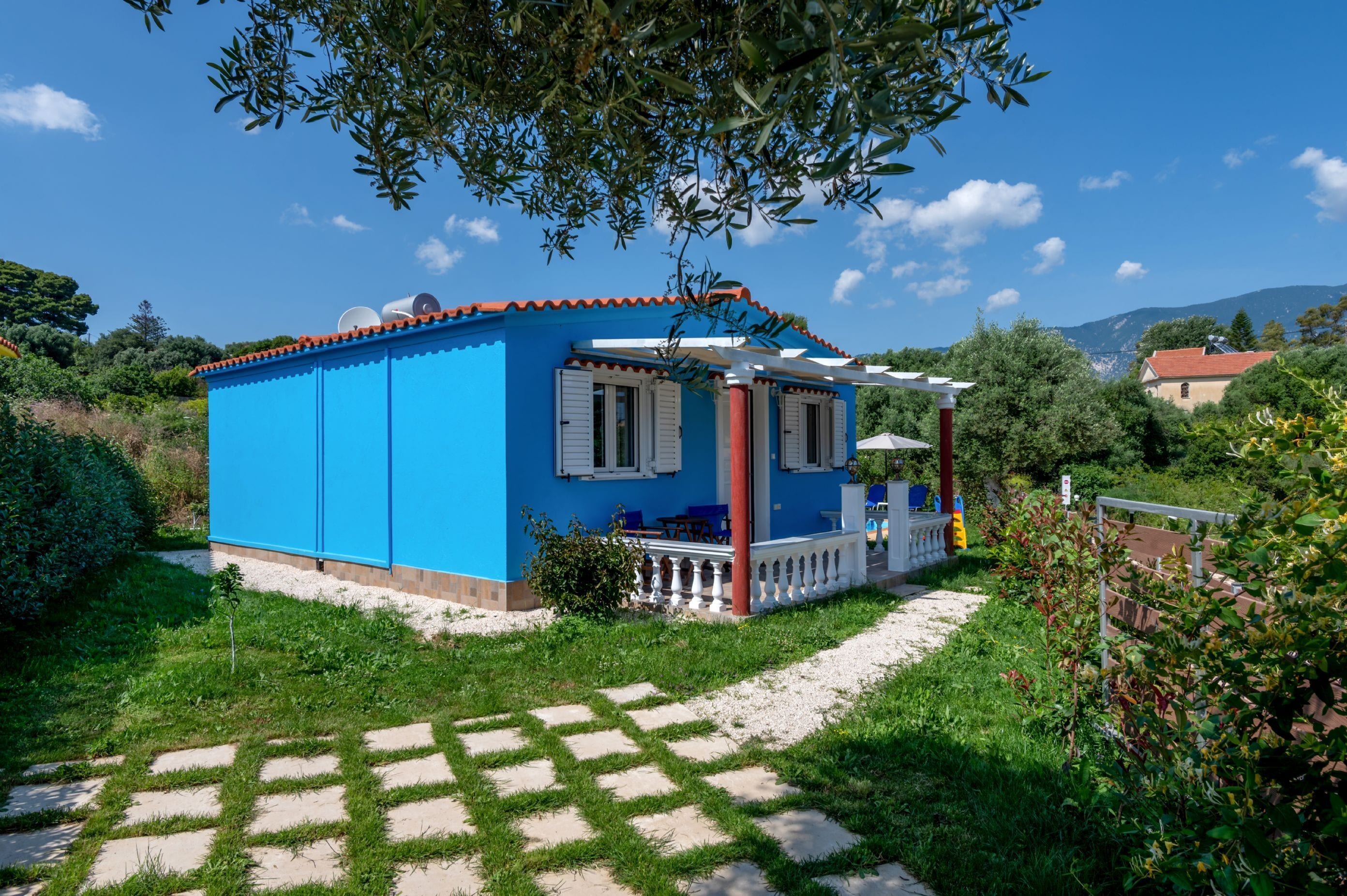 Une maison bleue avec un porche blanc, entourée de verdure et sous un ciel dégagé.
