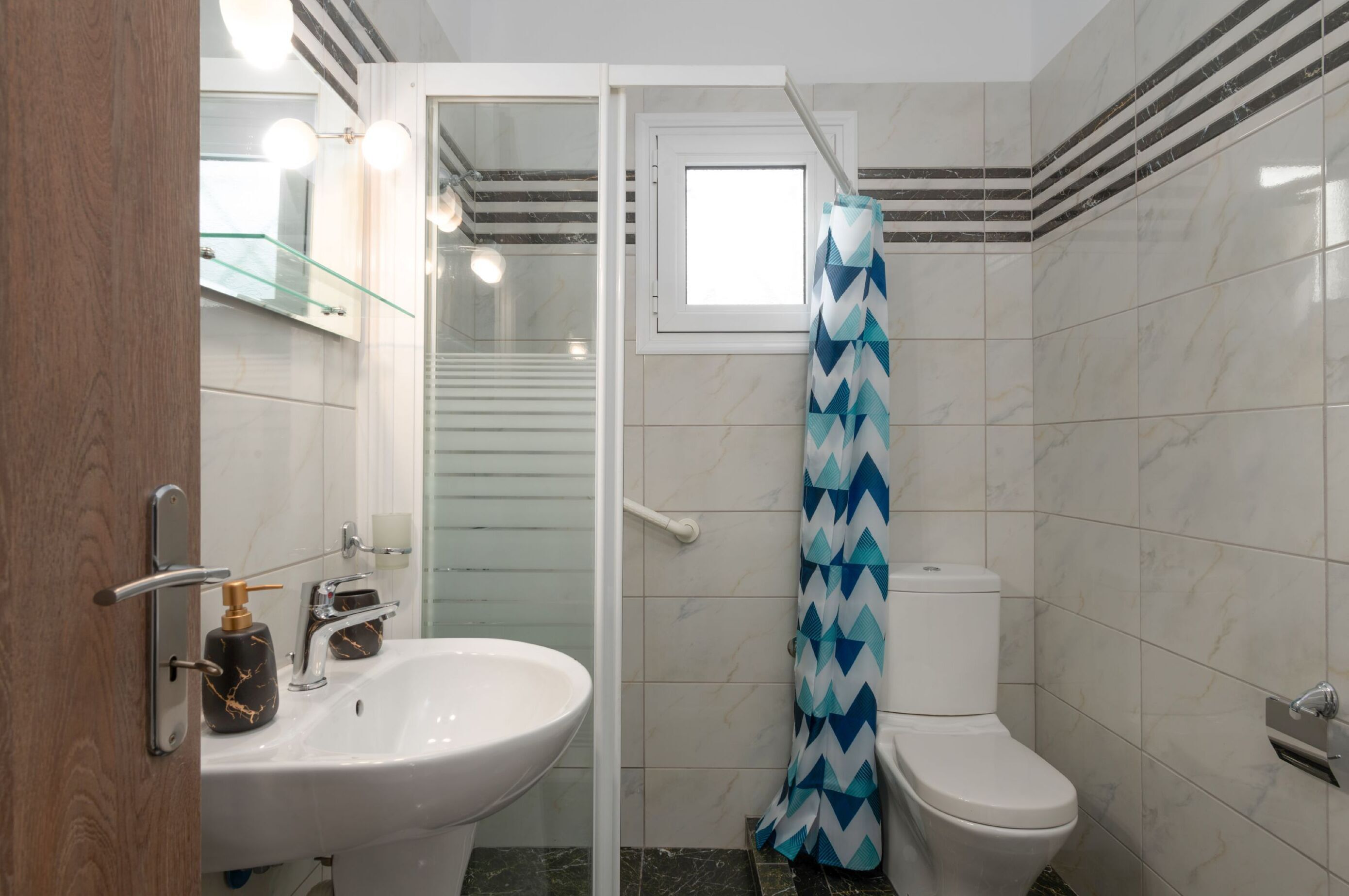 Une salle de bain moderne avec une douche à parois de verre, un lavabo blanc doté d’un distributeur de savon doré et des toilettes blanches.