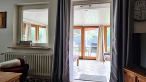 Een kamer met een houten kast, een rode stoel, een radiator, een raam met gordijnen en uitzicht op een buitenruimte met een tafel en stoelen.